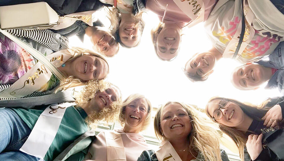 A hen party group smiling in a circle, looking down at the camera from above, all wearing sashes and casual outfits.