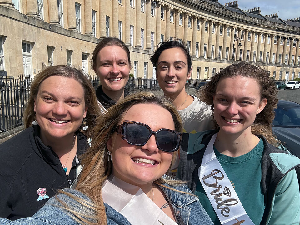 A group of women on a hen party taking a selfie, with bright sunshine and sashes reading “Bride Tribe” and “Bride to Be”.